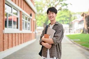 A cheerful Asian man student stands outside a brick building on campus, holding a laptop and books. photo