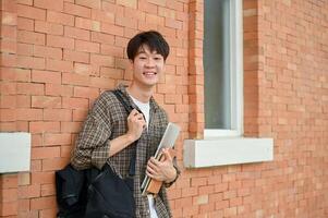 A smiling young Asian male college student stands by a brick wall on campus, carrying a backpack. photo