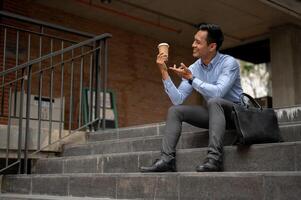 A handsome millennial Asian businessman enjoying his coffee on the stairs in front of the building. photo