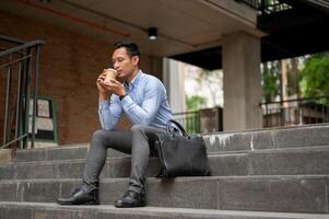 A handsome millennial Asian businessman enjoying his coffee on the stairs in front of the building. photo