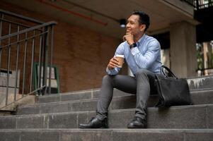 A handsome millennial Asian businessman enjoying his coffee on the stairs in front of the building. photo