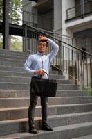 An exhausted Asian millennial businessman stands on the stairs, holding a coffee cup and a briefcase photo