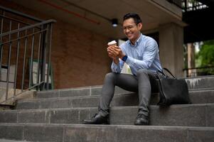 A handsome millennial Asian businessman enjoying his coffee on the stairs in front of the building. photo