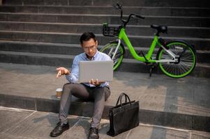 A confused Asian businessman sits on steps with a laptop, showing puzzled expression. photo