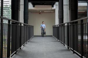 A handsome Asian businessman walking on a building corridor, holding a coffee cup and a briefcase. photo