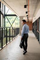 Determined Asian businessman walking along an indoor corridor, holding a coffee cup and a briefcase. photo