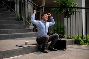 An angry Asian businessman sits on outdoor steps, holding a laptop with frustration, being fired. photo