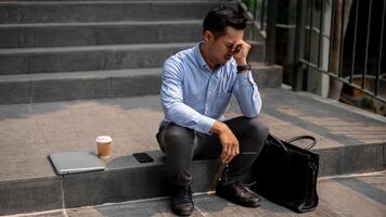 An Asian businessman sits on outdoor steps, experiencing frustration and the fear of being fired. photo