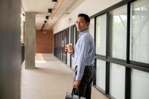 Determined Asian businessman walking along an indoor corridor, holding a coffee cup and a briefcase. photo