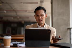 A confused Asian businessman is staring at his digital tablet screen with a puzzled expression. photo