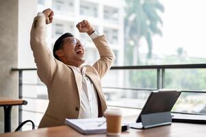 A cheerful Asian businessman raises his arms triumphantly, celebrating success or good news. photo