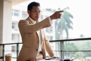 An angry Asian businessman is pointing his finger with angry expression, standing at a table. photo