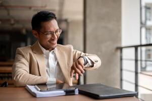 A confident Asian businessman checking time on his wristwatch while sitting in a building corridor. photo