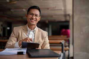 A confident Asian businessman sits at a table in a building corridor with his tablet and document. photo