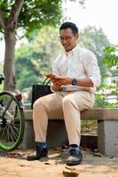 A happy Asian businessman sits on a bench in a public park, using his smartphone. photo