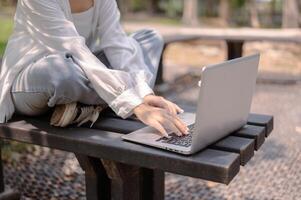 A cropped image of an Asian woman sits on a bench in a public park and uses her laptop computer. photo