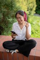 A cheerful Asian woman sits on a brick bench in a garden holding a phone with a raised fist gesture. photo