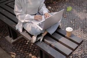 A cropped image of an Asian woman sits on a bench in a public park and uses her laptop computer. photo