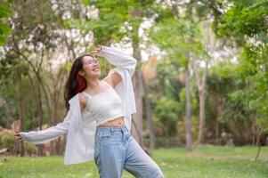 A young, happy Asian woman joyfully dances in a lush green park, expressing freedom. photo