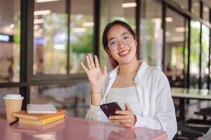 A female college student sits at a table in a campus cafeteria, smiling and waving at the camera. photo