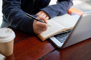 A woman is jotting down something in a notebook while working on her laptop at a table in a cafe. photo