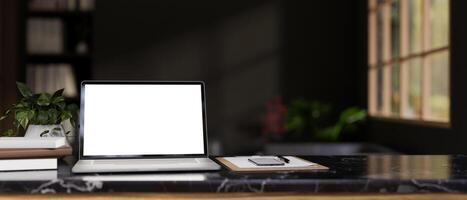 A modern workspace with a white-screen laptop mockup on a black marble table in a modern black room. photo