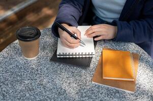 mujer tomando notas en un espiral bloc mientras sentado a un al aire libre mesa, trabajando remotamente a un café foto