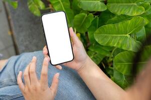 un mujer utilizando su teléfono inteligente mientras relajante en un verde jardín. un pantalla blanca teléfono inteligente Bosquejo. foto