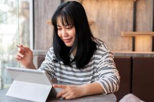 un contento asiático mujer es utilizando su digital tableta, trabajando remotamente a un café en el ciudad. foto