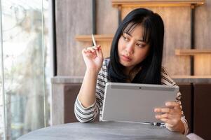 un grave asiático mujer es utilizando su digital tableta, pensando y planificación su trabajo en su cabeza. foto