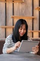 un contento asiático mujer es utilizando su digital tableta, trabajando remotamente a un café en el ciudad. foto