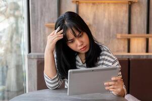 un estresado asiático mujer mira a su tableta pantalla con un grave rostro, contempla. foto