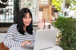 A positive Asian woman is having an online meeting with her team, working remotely at a cafe. photo