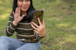 A happy Asian woman is talking on a call with her friend while relaxing in a green park. photo