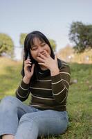A cheerful young Asian woman enjoying talking on the phone with her friend while relaxing in a park. photo