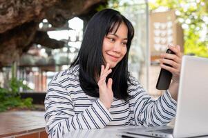 A positive Asian woman is talking on a call on her phone while working remotely at a cafe. photo