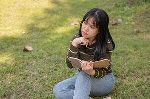 A young Asian woman is thinking about some ideas while doing her homework, sitting in a park. photo