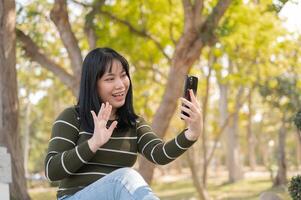 A cheerful Asian woman is talking on a call with her friend while relaxing in a green park. photo