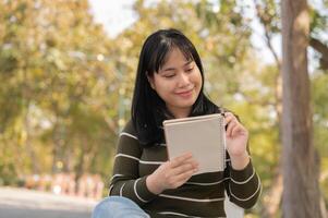 un bonito asiático mujer es sentado en un banco en un verde parque con su computadora portátil, acuerdo su diario. foto