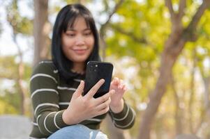 A positive young Asian woman using her smartphone while relaxing on a bench in a green park. photo