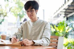 A happy Asian man is focusing on writing in his notebook while sitting at an outdoor table in a cafe photo