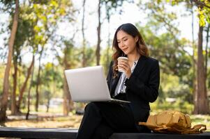 A beautiful Asian businesswoman is sitting on a bench in a city park, and working on her laptop. photo