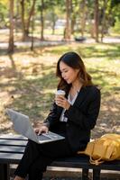 A beautiful Asian businesswoman is sitting on a bench in a city park, and working on her laptop. photo
