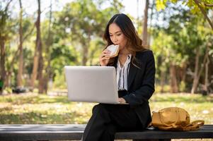 un ocupado, elegante asiático mujer de negocios es trabajando remotamente en su ordenador portátil en un parque mientras teniendo almuerzo. foto