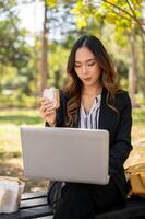 un ocupado, elegante asiático mujer de negocios es trabajando remotamente en su ordenador portátil en un parque mientras teniendo almuerzo. foto