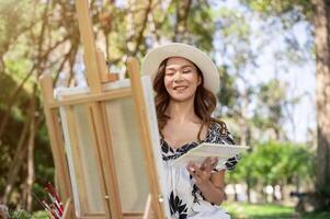 An attractive Asian female freelance artist is painting a picture on a canvas easel in a green park. photo