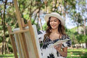 A charming Asian woman is painting a picture on a canvas easel while picnicking in a green park. photo