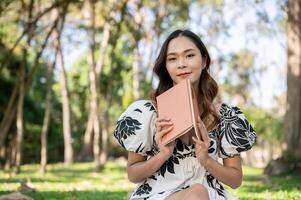 A beautiful Asian woman in a cute dress is reading a book in a green park on a bright, sunny day. photo