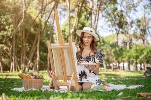 A charming Asian woman is painting a picture on a canvas easel while picnicking in a green park. photo