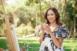 Positive Asian woman is enjoying hot tea and painting on a canvas easel while picnicking in a park. photo